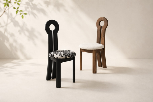 Two modern sculptural dining chairs with circular cutout backs, shown on a warm beige studio floor with soft leaf shadows. One chair is black with a black-and-white patterned seat cushion, and the other is medium brown wood with a white cushion.