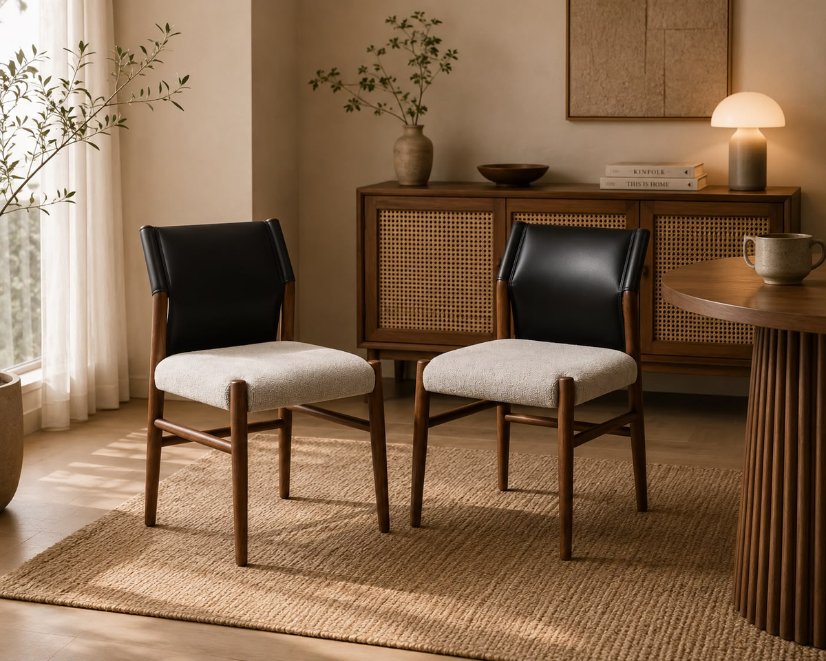 Two modern dining chairs with black leather backs and light beige upholstered seats, placed in a warm, sunlit room with a wooden cabinet, rug, and table in the background.