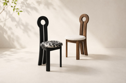 Two modern sculptural dining chairs with circular cutout backs, shown on a warm beige studio floor with soft leaf shadows. One chair is black with a black-and-white patterned seat cushion, and the other is medium brown wood with a white cushion.
