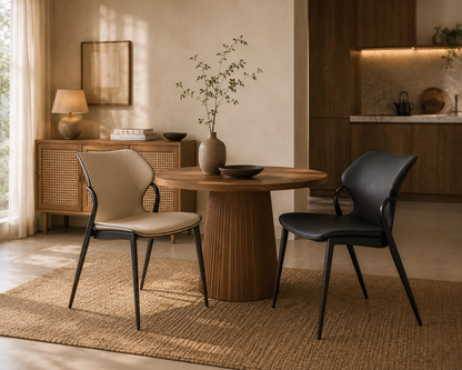 A styled dining room scene featuring two modern accent chairs—a beige upholstered chair with black legs and a black leather chair—around a round wooden table on a woven rug, with a sideboard and warm-toned interior in the background.